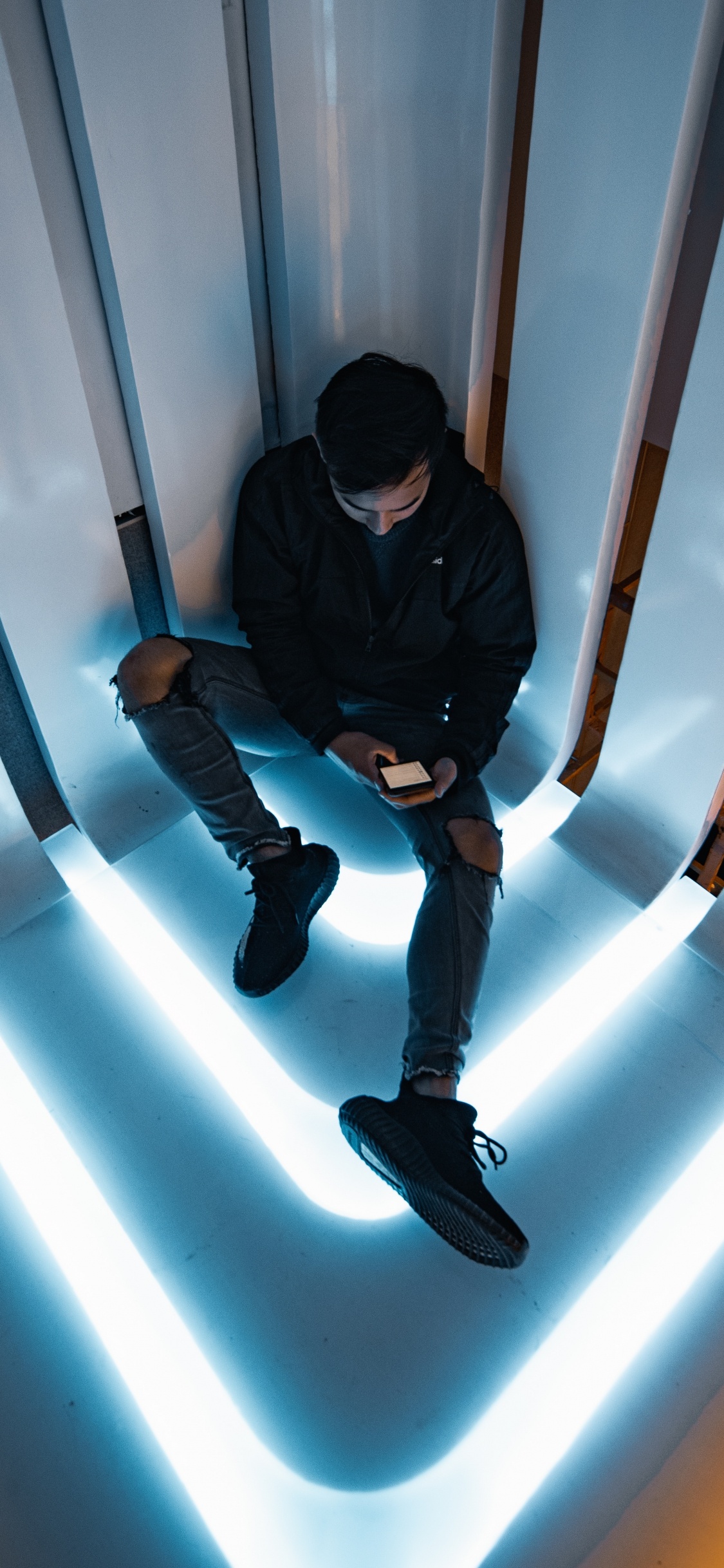 Man in Black Jacket and Black Pants Sitting on White Wall. Wallpaper in 1125x2436 Resolution