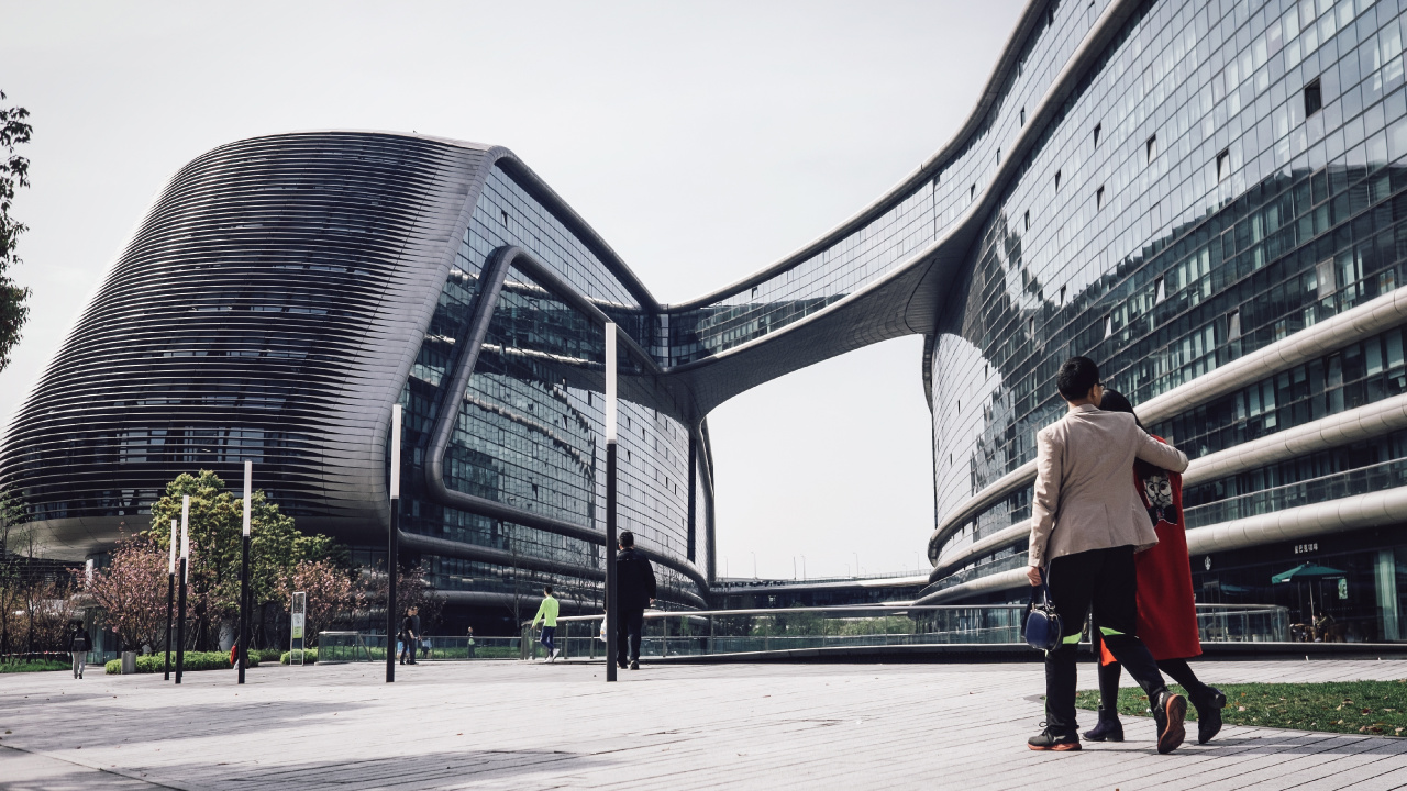 Woman in White Long Sleeve Shirt and Black Pants Walking on Sidewalk Near Glass Building During. Wallpaper in 1280x720 Resolution