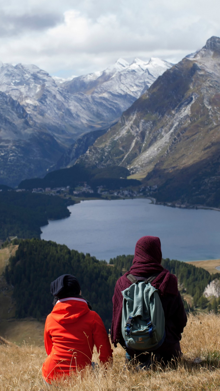Person in Red and Black Hoodie Sitting on Green Grass Field Near Lake and Mountains During. Wallpaper in 750x1334 Resolution