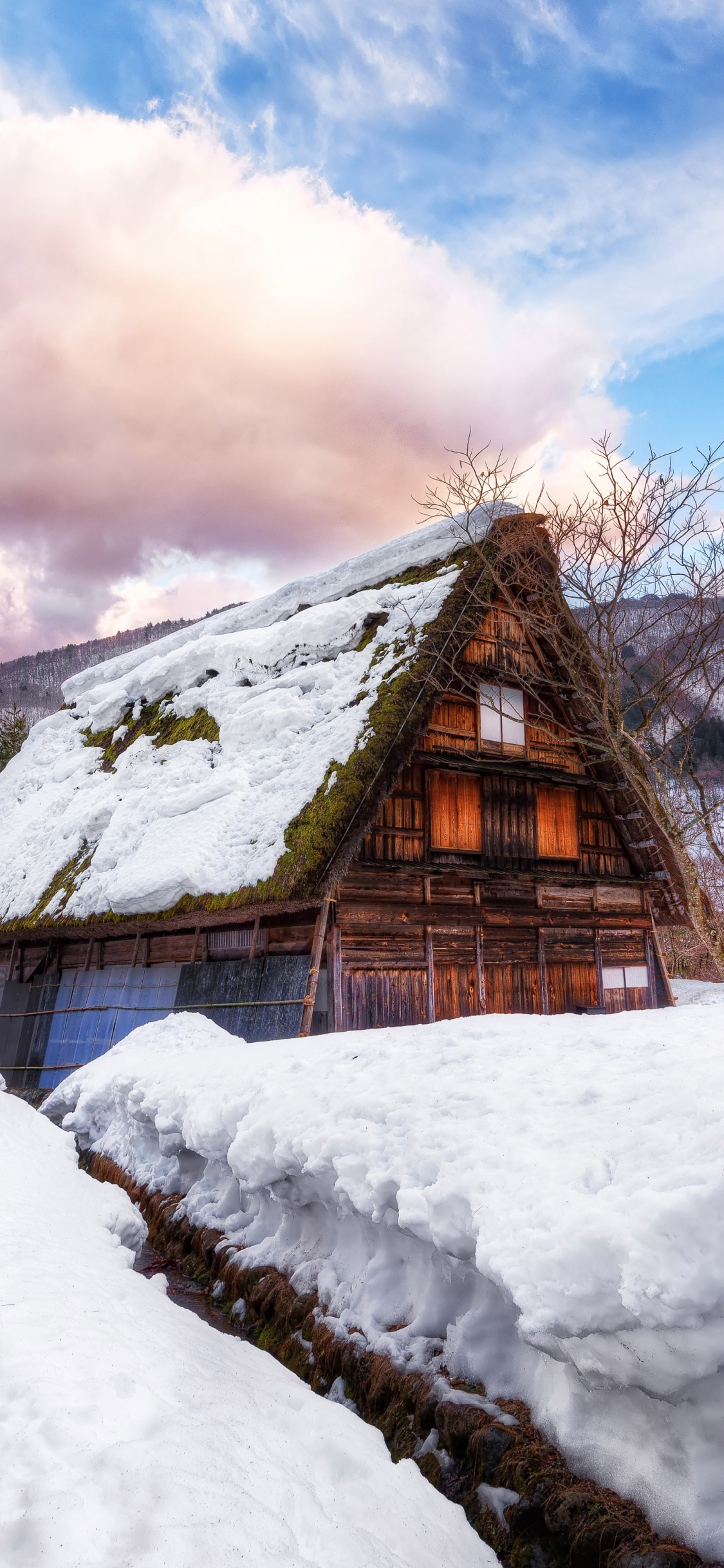 Casa de Madera Marrón Sobre Suelo Cubierto de Nieve Bajo Las Nubes Blancas y el Cielo Azul Durante el Día. Wallpaper in 1125x2436 Resolution