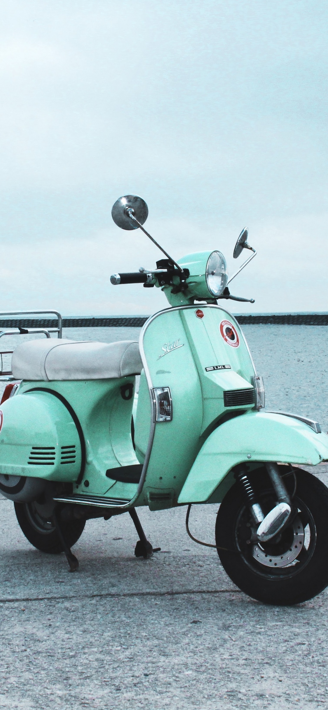 Green Motor Scooter Parked on Gray Concrete Pavement Near Body of Water During Daytime. Wallpaper in 1125x2436 Resolution