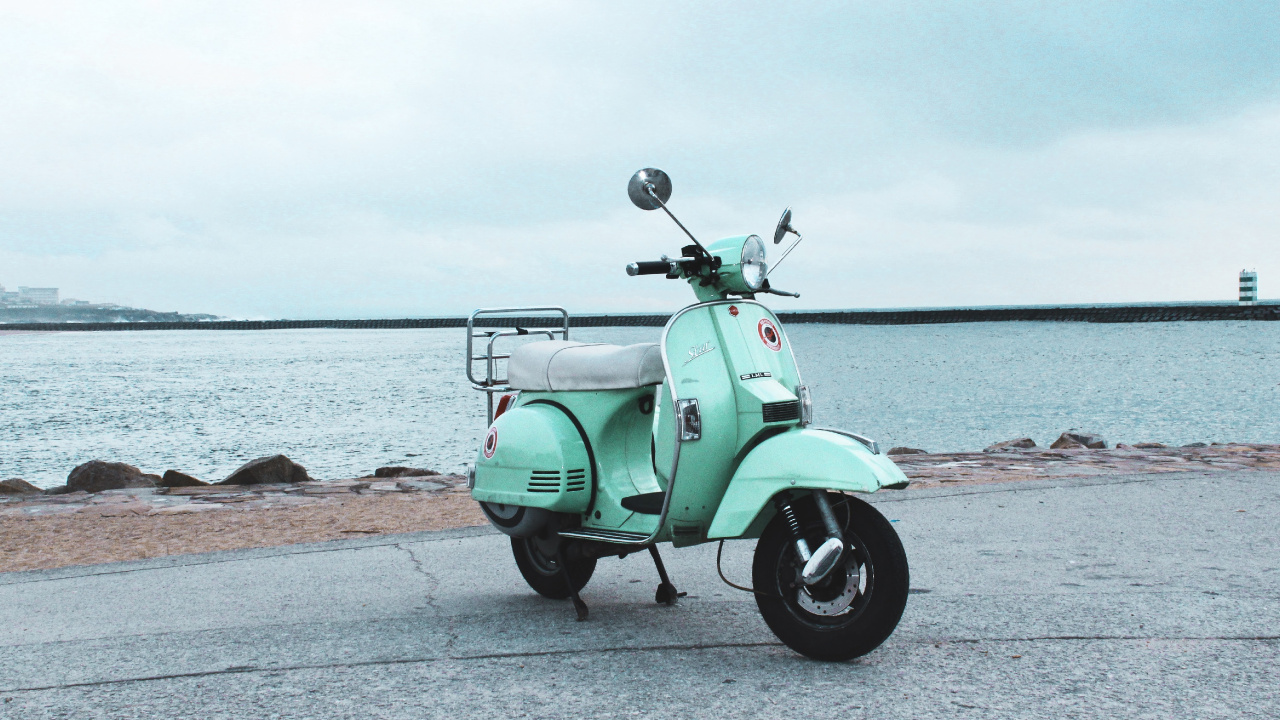 Green Motor Scooter Parked on Gray Concrete Pavement Near Body of Water During Daytime. Wallpaper in 1280x720 Resolution