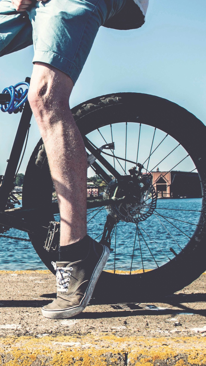 Person in Blauen Jeans Und Schwarzen Stiefeln, Die Ein Schwarzes Mountainbike in Der Nähe Eines Gewässers Fährt. Wallpaper in 720x1280 Resolution