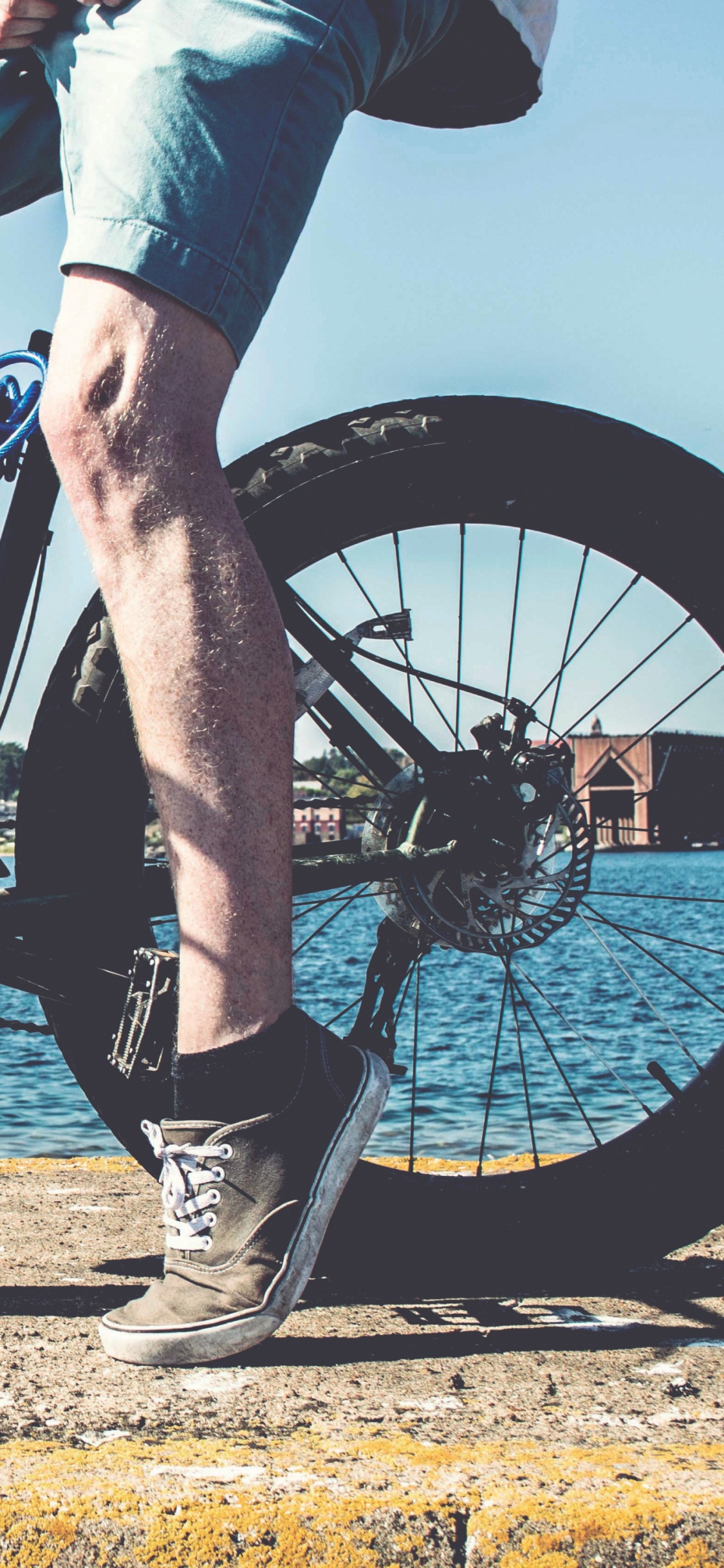 Persona en Jeans Azul y Botas Negras Montando Bicicleta de Montaña Negra Cerca Del Cuerpo de Agua. Wallpaper in 1125x2436 Resolution