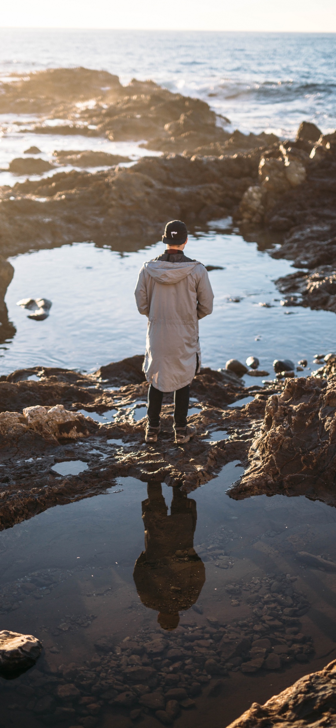 Man in White Jacket Standing on Rock Near Body of Water During Daytime. Wallpaper in 1125x2436 Resolution