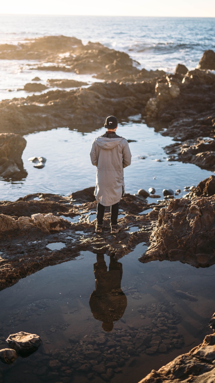 Man in White Jacket Standing on Rock Near Body of Water During Daytime. Wallpaper in 720x1280 Resolution