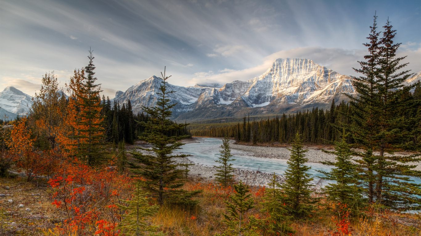 Green and Brown Trees Near Snow Covered Mountain Under Cloudy Sky During Daytime. Wallpaper in 1366x768 Resolution