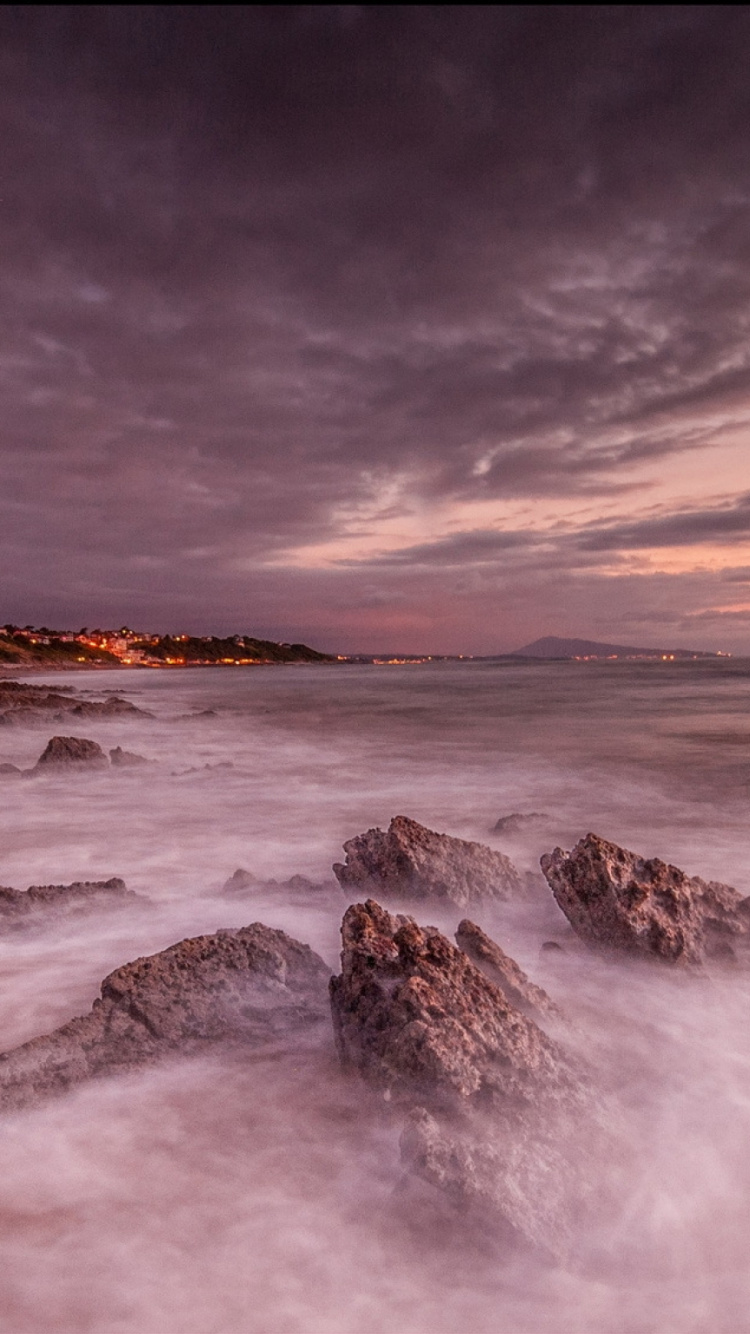 Olas Del Océano Rompiendo Sobre Rocas Bajo Nubes Grises. Wallpaper in 750x1334 Resolution