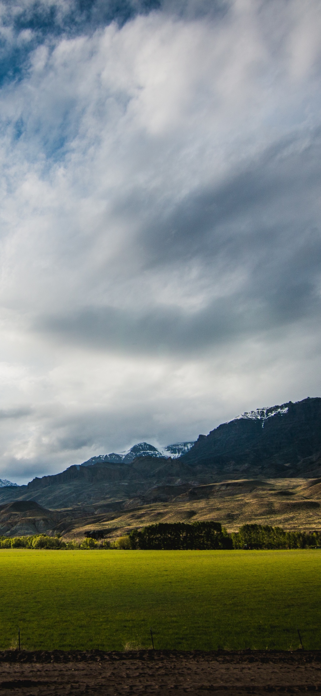 Green Grass Field Near Brown Mountain Under White Clouds and Blue Sky During Daytime. Wallpaper in 1125x2436 Resolution