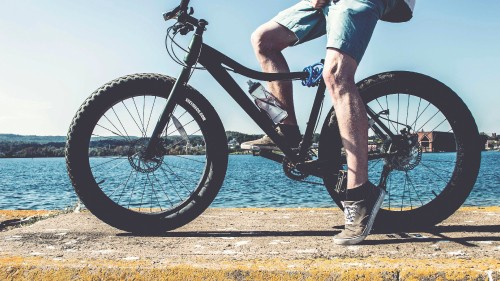 Image person in blue denim jeans and black boots riding black mountain bike near body of water