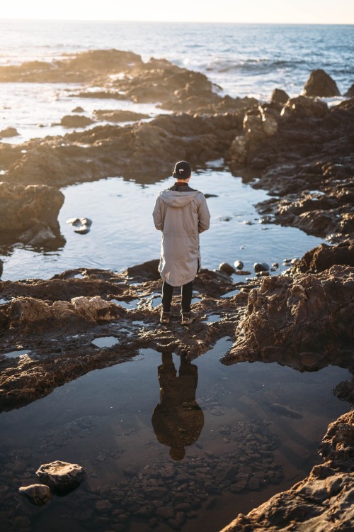 Image man in white jacket standing on rock near body of water during daytime