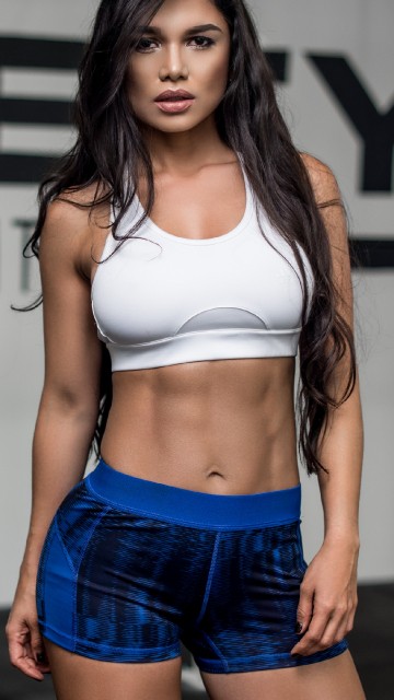 Image woman in white sports bra and blue denim shorts standing near white wall